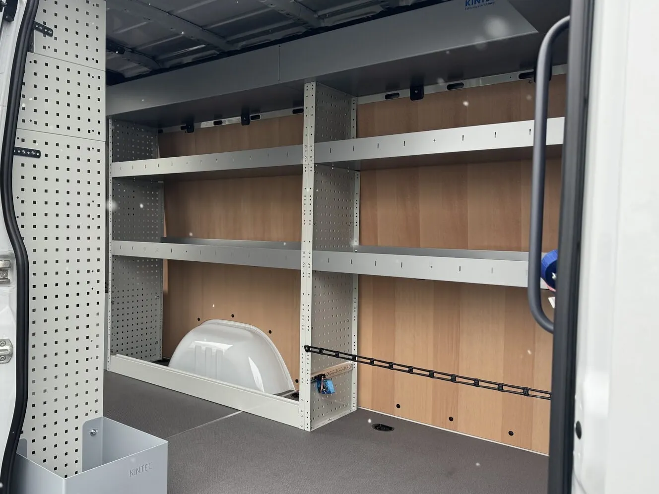 Regalzeile mit durchgehenden Fachböden über dem Radkasten, dahinter eine Sperrholzwand, unten ein schwarzes Spannband